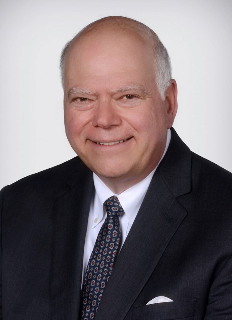 This image shows a smiling person with a bald head, wearing a dark suit, patterned tie, and a white pocket square, against a white background.